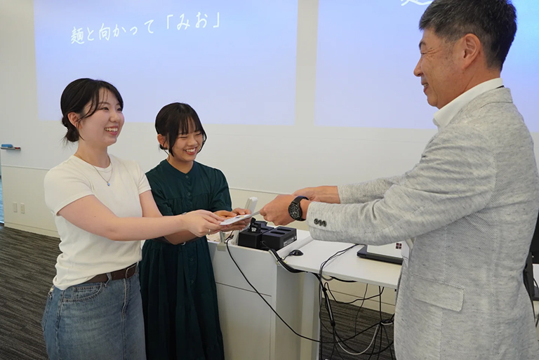 麺と向かって「みお」 - 「地域×学生で挑む、ご当地ラーメンを活かした観光地域活性化」 「すでに活動をしているプロジェクトの新規事業の展開」として注目を集めました。実店舗での販売実績を基盤に、お土産用ラーメンの開発やふるさと納税への返礼品提案など、地域貢献と美浜町全体への利点に繋がる点が評価されています。地元政府や地域関係者との連携も強みであり、将来の学生がビジネス運営や地域との協働を学ぶ機会となる点も期待されています。